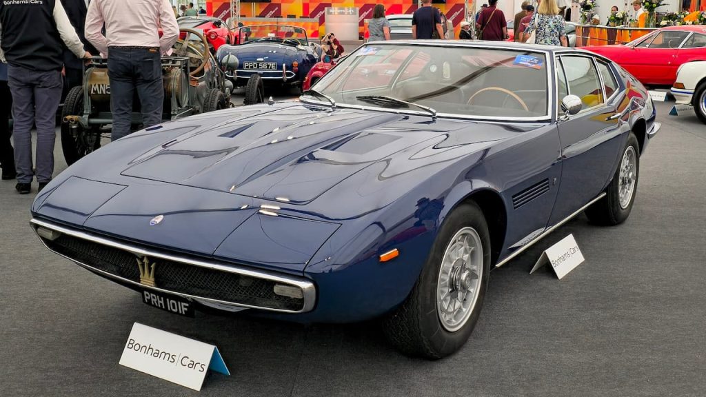 Dark blue 1968 Maserati Ghibli with registration PRH101F displayed at Bonhams Cars auction with visitors and classic cars nearby
