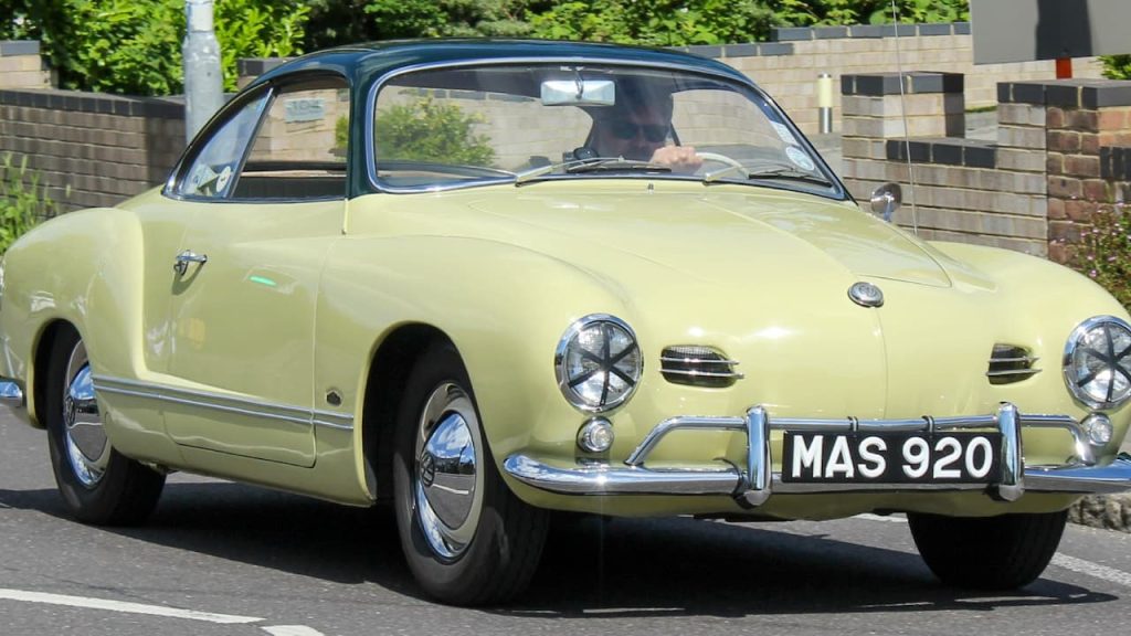 Pale yellow Volkswagen Karmann Ghia with black roof and registration MAS920 driving on street with brick buildings behind
