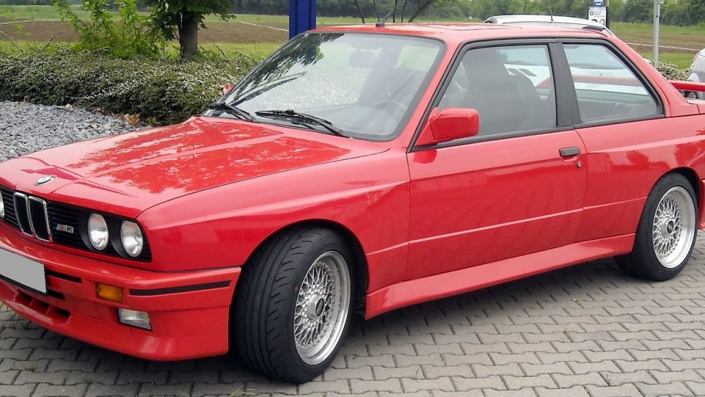 Red BMW M3 E30 classic sports car with wire-spoke wheels and M3 badge parked on cobblestone surface