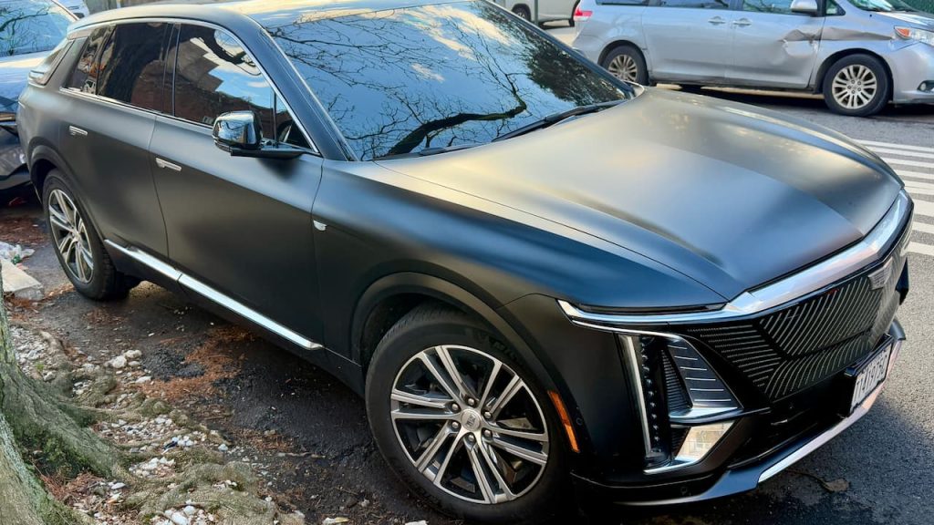 Dark gray Cadillac Lyriq electric SUV with distinctive front grille and multi-spoke wheels parked on street